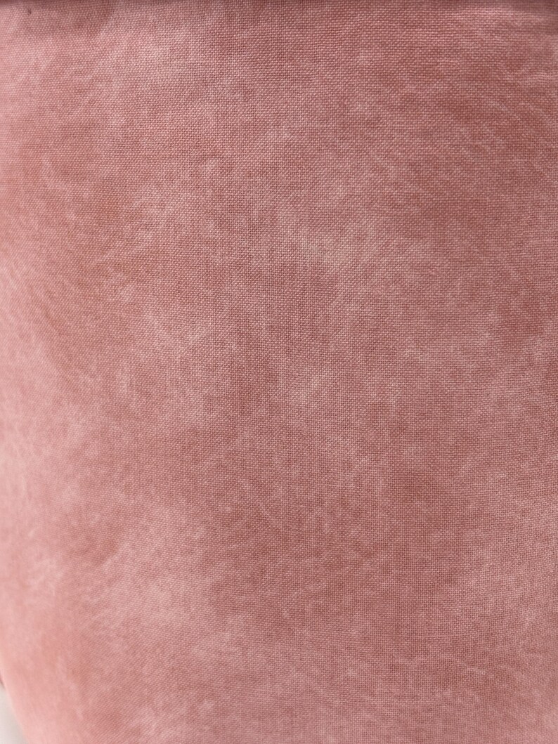 May include: Close-up of a dusty rose fabric with a textured appearance. The material has a slightly faded look, with lighter areas creating a subtle pattern across the surface. The fabric appears to be a soft, woven textile.