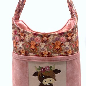 May include: A shoulder bag with a pink suede-like exterior and a floral patterned interior. The bag features a large embroidered image of a Highland cow wearing a flower crown. The bag's strap is made of the same floral fabric as the interior.