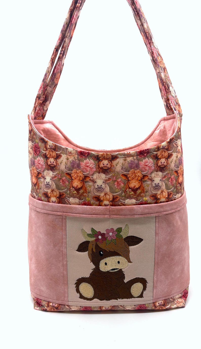 May include: A shoulder bag with a pink suede-like exterior and a floral patterned interior. The bag features a large embroidered image of a Highland cow wearing a flower crown. The bag's strap is made of the same floral fabric as the interior.