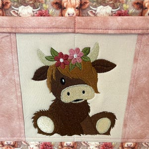 May include: A pink and cream-colored fabric item featuring an embroidered Highland cow wearing a floral crown. The cow is brown with cream-colored accents. The background has a cow print.