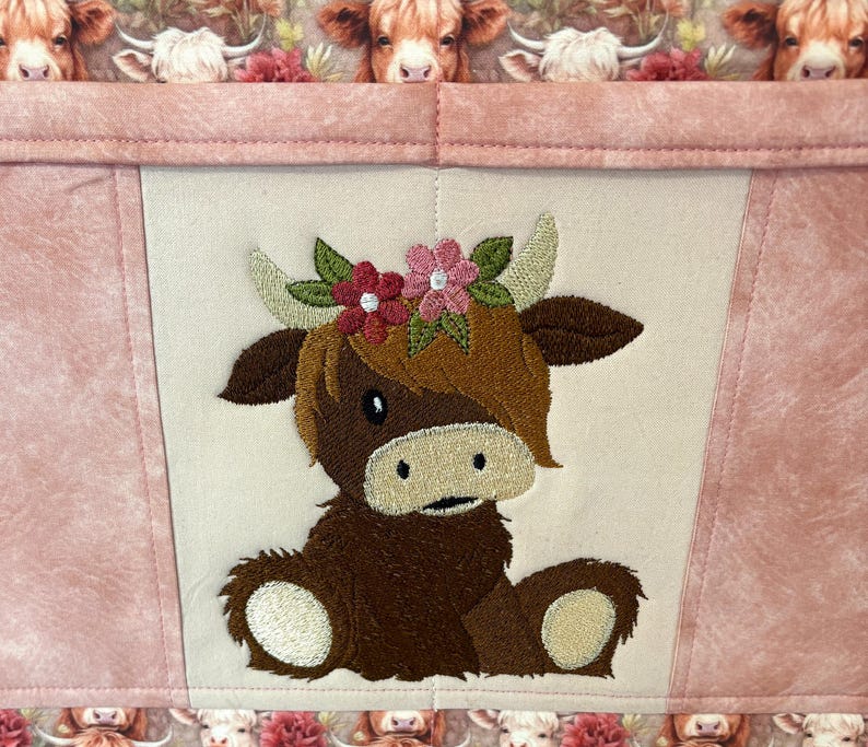 May include: A pink and cream-colored fabric item featuring an embroidered Highland cow wearing a floral crown. The cow is brown with cream-colored accents. The background has a cow print.
