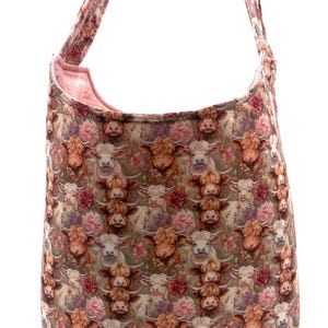 May include: A large shoulder bag with a repeating pattern of Highland cows and pink flowers. The bag has a light pink interior and matching straps. The bag is made of a soft fabric.