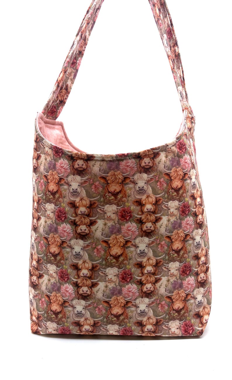 May include: A large shoulder bag with a repeating pattern of Highland cows and pink flowers. The bag has a light pink interior and matching straps. The bag is made of a soft fabric.