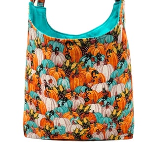 May include: A turquoise blue tote bag with a pumpkin print. The print features orange, white, and teal pumpkins with autumn foliage and corn.