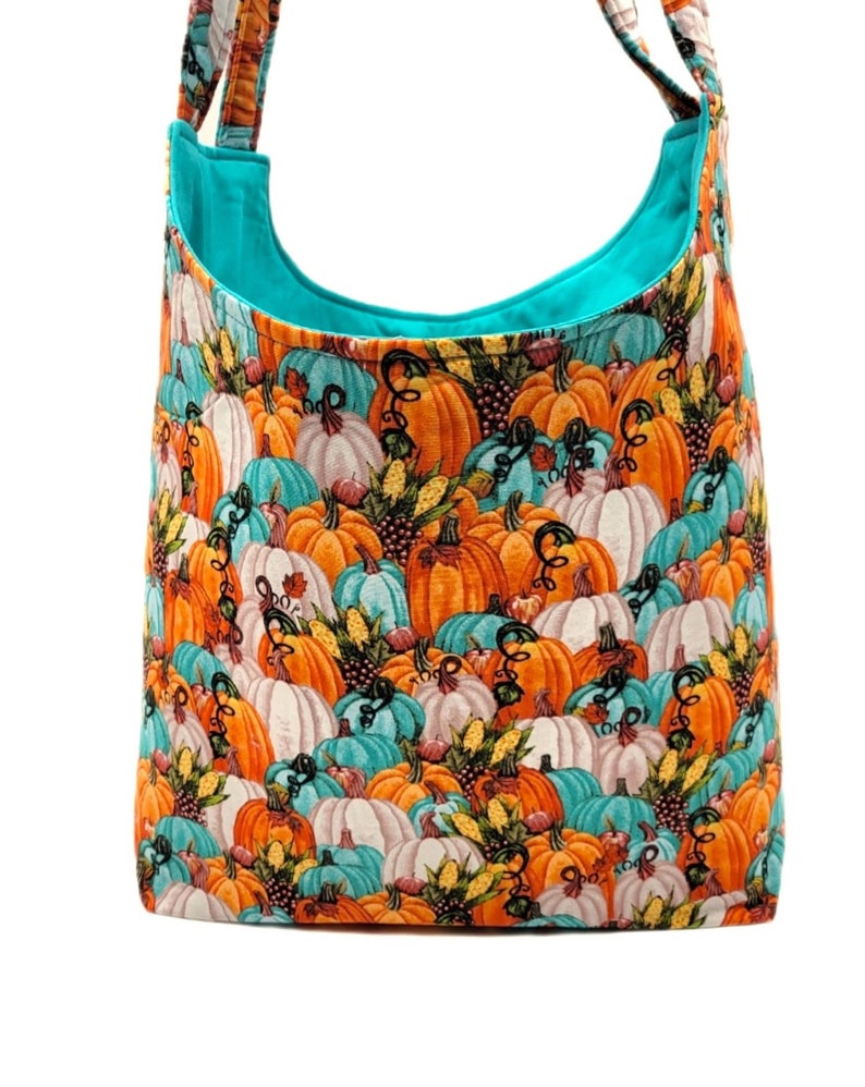 May include: A turquoise blue tote bag with a pumpkin print. The print features orange, white, and teal pumpkins with autumn foliage and corn.