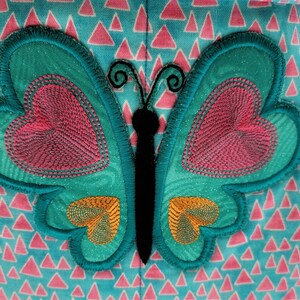 May include: A teal butterfly applique with pink heart-shaped details and orange accents. The butterfly has a black body and antennae. The background is a light blue fabric with pink triangle patterns.