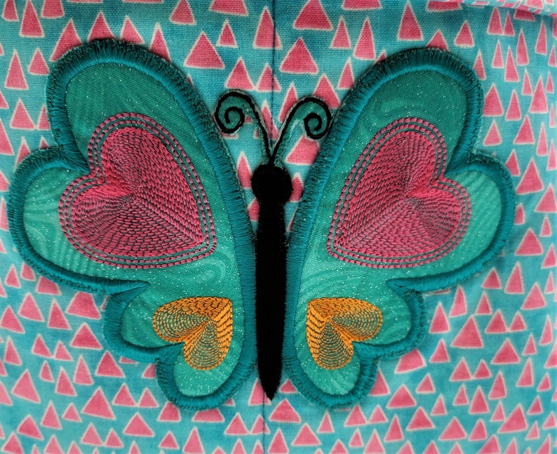 May include: A teal butterfly applique with pink heart-shaped details and orange accents. The butterfly has a black body and antennae. The background is a light blue fabric with pink triangle patterns.