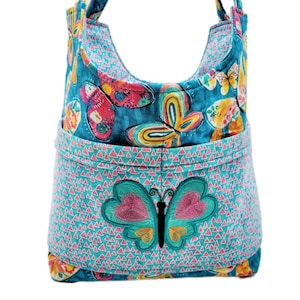 May include: A turquoise fabric tote bag with a butterfly design. The bag features a large embroidered butterfly on the front pocket and a repeating butterfly pattern. The bag has a curved top and two handles.