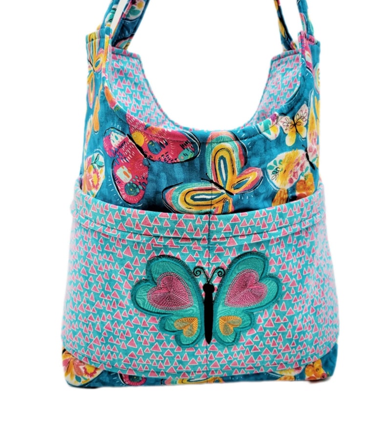 May include: A turquoise fabric tote bag with a butterfly design. The bag features a large embroidered butterfly on the front pocket and a repeating butterfly pattern. The bag has a curved top and two handles.