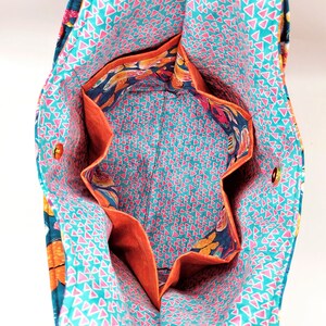May include: Open view of a fabric tote bag with multiple interior pockets. The bag's interior features a teal background with pink triangles, while the pockets are lined with orange fabric. The exterior has a floral pattern.