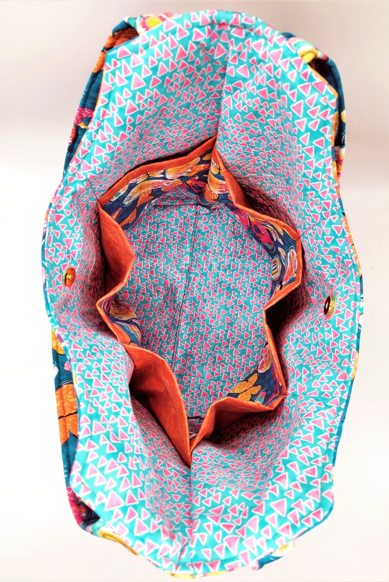 May include: Open view of a fabric tote bag with multiple interior pockets. The bag's interior features a teal background with pink triangles, while the pockets are lined with orange fabric. The exterior has a floral pattern.