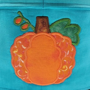 May include: A bright orange pumpkin with a brown stem and green leaves is appliqued on a turquoise fabric background.