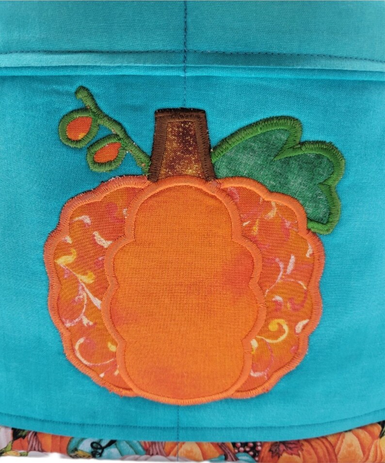 May include: A bright orange pumpkin with a brown stem and green leaves is appliqued on a turquoise fabric background.
