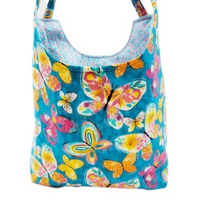 May include: A turquoise tote bag with a colorful butterfly pattern. The butterflies are in shades of pink, yellow, and orange, with a white background. The bag has a contrasting pink and blue patterned lining and matching straps.