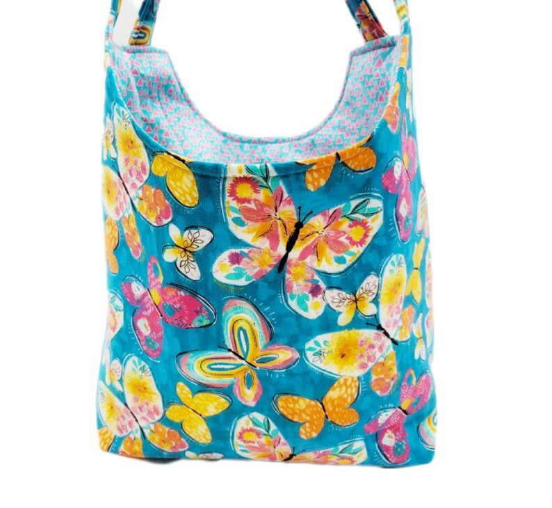 May include: A turquoise tote bag with a colorful butterfly pattern. The butterflies are in shades of pink, yellow, and orange, with a white background. The bag has a contrasting pink and blue patterned lining and matching straps.