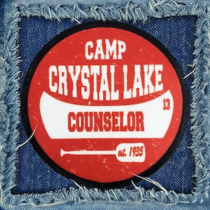 May include: Red and white circular patch with the text "Camp Crystal Lake Counselor est. 1935" and a canoe graphic.