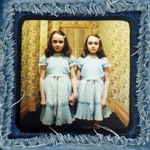 May include: Two girls in matching blue dresses stand in a hallway with floral wallpaper. They are holding hands and looking at the camera.