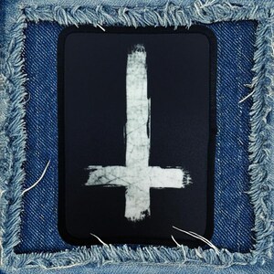 May include: A black patch with a white upside-down cross painted on it. The cross is made with a brushstroke effect.