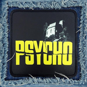 May include: A black and yellow patch with the word "PSYCHO" in large yellow letters. The patch is sewn onto a blue denim background. A black and white image of a house is behind the text.
