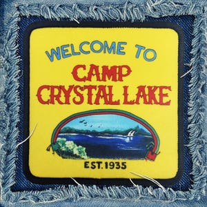 May include: A yellow patch with the text "Welcome to Camp Crystal Lake Est. 1935" in a blue denim background.