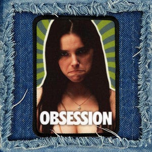 May include: A rectangular fabric patch featuring a photographic print of a person with long dark hair, a thin necklace, and a serious expression. The background has green and blue radiating lines. The word OBSESSION is printed in white at the bottom.