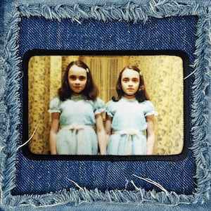 May include: A denim patch with a black border featuring a vintage photograph of two girls in blue dresses standing in a room with wallpaper.