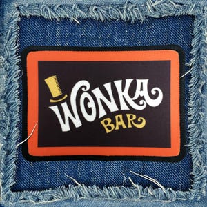 May include: A black and orange fabric patch with a white Wonka Bar logo and a yellow top hat graphic. The text "Wonka Bar" is in a stylized font.