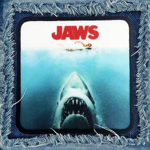May include: A blue denim patch with a white background and the word "JAWS" in red letters. The image shows a shark's open mouth with sharp teeth, partially submerged in blue water.