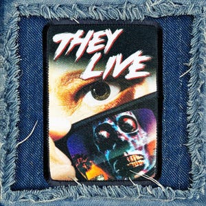 May include: A black and white patch with the words "They Live" in red and white letters. The patch features a close-up of an eye with a distorted image of an alien face behind it.