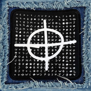 May include: A black and white patch with a circle and cross design on a blue denim background. The circle and cross are surrounded by a grid of letters and symbols.