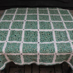 May include: A handmade crochet blanket featuring a grid pattern of square motifs. The squares are a variegated mix of light and dark green yarn, separated by a solid off-white yarn border that creates a distinct lattice effect across the fabric.