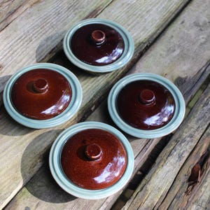 May include: Four brown ceramic bowls with light blue rims and lids. The bowls are arranged in a square pattern on a wooden surface.