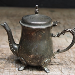 May include: A tarnished silver-toned metal teapot featuring a curved spout, an ornate handle, and a lid topped with a small finial. The teapot stands on three decorative feet and displays a weathered, dark patina across its entire surface.
