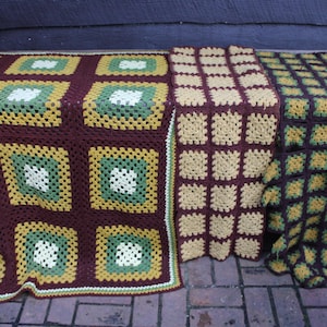 May include: Three crocheted blankets featuring granny square patterns. The first blanket has large squares in dark brown, olive green, and cream. The second is a solid cream and brown grid. The third features small squares in dark brown and yellow.