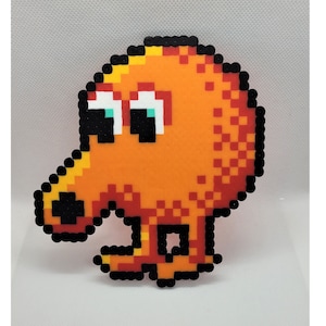 May include: A pixel art character made of orange, black, and white beads. The character has a round body, two eyes, and a beak-like nose. It has two short legs.