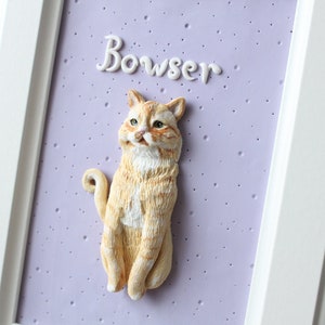 May include: A white frame with a light purple background and a white cat figurine. The cat is sitting up with its tail curled around its legs. The cat is orange and white with a white patch on its chest. The name "Bowser" is written in white cursive above the cat.