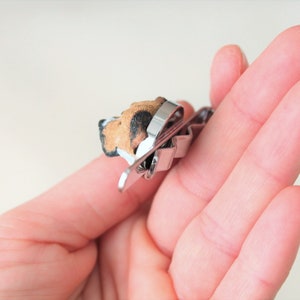 May include: A silver metal tie clip with a small, brown and black dog figurine attached. The dog is facing to the left and has a white patch on its chest.