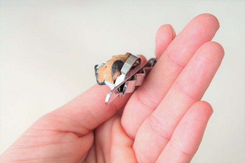 May include: A silver metal tie clip with a small, brown and black dog figurine attached. The dog is facing to the left and has a white patch on its chest.