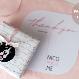 May include: A white gift box with a black and white dog illustration on a pink background. The box is tied with a pink ribbon. A white card with the text "Thank you" and "NICO MADE .ME" is on the right side of the image. The text "Packaging sample" is on the top right corner of the image.