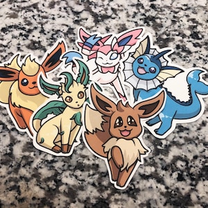 Op de afbeelding: Vijf kleurrijke cartoon Pokemon-stickers met Eevee, Vaporeon, Flareon, Jolteon en Espeon. De stickers zijn gedrukt op wit papier en hebben een glanzende afwerking.