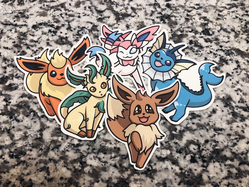 Op de afbeelding: Vijf kleurrijke cartoon Pokemon-stickers met Eevee, Vaporeon, Flareon, Jolteon en Espeon. De stickers zijn gedrukt op wit papier en hebben een glanzende afwerking.
