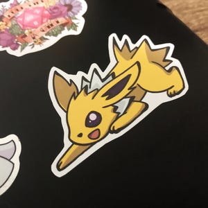 Op de afbeelding: Een gele en witte cartoon sticker van een Pokemon-personage met stekelig haar en een grote glimlach. Het personage staat op een zwarte achtergrond.