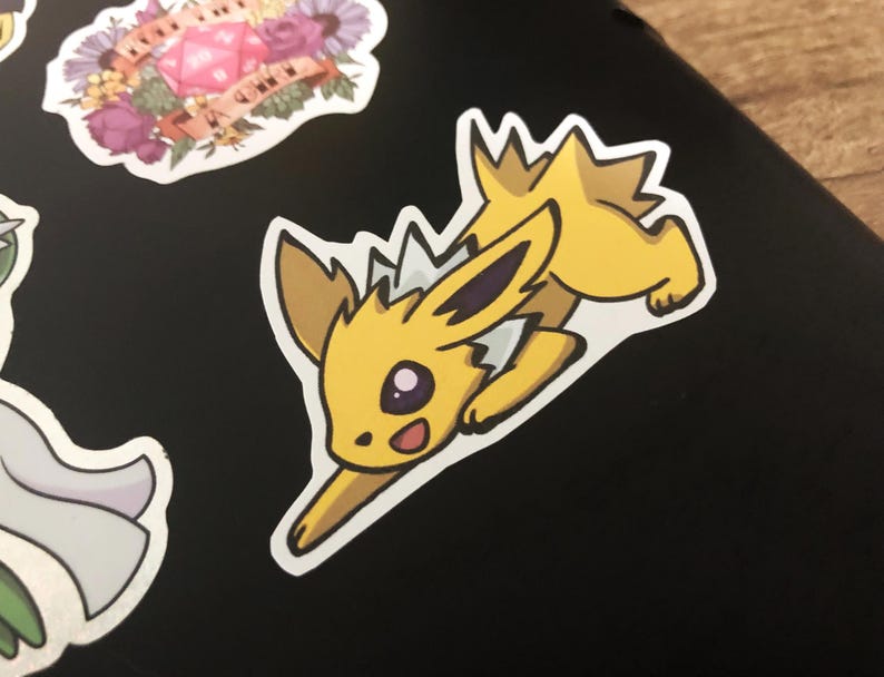 Op de afbeelding: Een gele en witte cartoon sticker van een Pokemon-personage met stekelig haar en een grote glimlach. Het personage staat op een zwarte achtergrond.