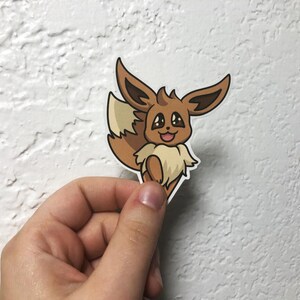 Op de afbeelding: Een bruine en beige cartoon Eevee sticker met grote ogen en een vrolijke uitdrukking. De sticker is op een witte achtergrond.