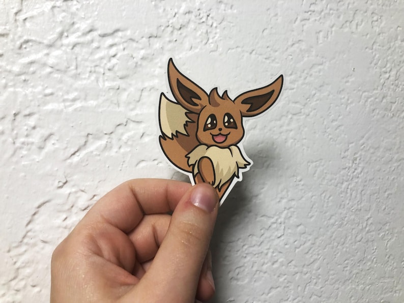 Op de afbeelding: Een bruine en beige cartoon Eevee sticker met grote ogen en een vrolijke uitdrukking. De sticker is op een witte achtergrond.