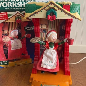 May include: Animated Christmas puppet theater featuring Mrs. Claus holding a cookie and a mug. The red and white figure is in a red and yellow structure with the text "SANTA'S COOKIE BREAK". The box reads "SANTA'S NORTH POLE WORKSHOP".