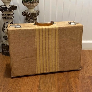 May include: A rectangular tan suitcase with a textured fabric exterior featuring a vertical striped panel in the center. The luggage includes a curved amber-colored plastic handle and two silver-toned metal latches on the top edge for closure.