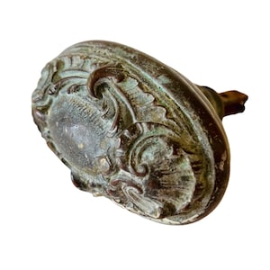 May include: An antique oval-shaped door knob with intricate floral designs. The knob is made of metal and has a greenish patina.