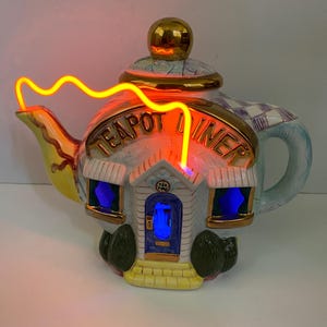 May include: A white ceramic teapot shaped like a diner with a neon sign that reads "Teapot Diner". The teapot has a blue door and windows with a yellow base and green bushes.