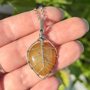 May include: A teardrop-shaped orange gemstone pendant wrapped in silver-colored wire arranged in a leaf-like pattern. The pendant is attached to a silver-colored metal chain link necklace, featuring a coiled wire bail at the top of the stone.